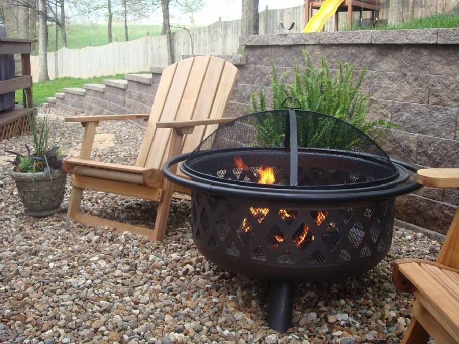 A backyard with a round, mesh-covered fire pit, wooden Adirondack chairs, and potted plants on a pebble ground, with a wooden playset in the distance.